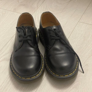 Svarta Dr. Martens skor med gula sömmar - Klassiska äkta Dr. Martens i skinn med rund tå och tydliga gula sömmar runt sulan. Skorna har snörning och en robust platt sula som ger en ikonisk look. Knappt använda, kanske runt 3-4 gånger så praktiskt taget nyskick. Storlek 37. 