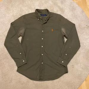 Ralph Lauren skjorta - Ralph Lauren Skjorta – Slim Fit – M  Säljer en snygg och klassisk skjorta från Ralph Lauren i färgen olivgrön. Modellen är Slim Fit och storlek Medium (M). Ikonisk orange Polo-logga broderad på bröstet. Skjortan är tillverkad i ett mjukt och slitstarkt bomullstyg och passar perfekt till både vardag och mer uppklädda tillfällen.  ✅ Bra skick ✅ Knappar i full funktion ✅ Tillverkad i Filippinerna ✅ 100 % äkta Ralph Lauren