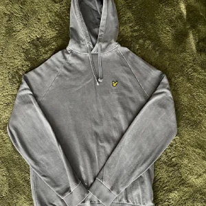 Grå hoodie från Lyle & Scott - Säljer en stilren grå hoodie från Lyle & Scott med klassisk huva och dragsko. Tröjan har ett litet broderat gult logomärke på bröstet och ribbade muddar vid ärmslut och nederkant. Perfekt för en avslappnad look.
