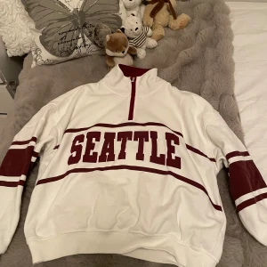 Vit sweatshirt med röd text från STAY - Säljer en vit sweatshirt från STAY med röd text 'SEATTLE' och röda detaljer på ärmarna. Tröjan har halv dragkedja vid halsen och ribbade muddar. Perfekt för en avslappnad stil.