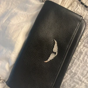 Zadig & Voltaire väska - Säljer en svart axelväska i skinnimitation med silverfärgade detaljer och kedjerem. Väskan har en dekorativ silverlogga framtill och dragkedja på baksidan. Perfekt storlek för det viktigaste och stilren design. Saknar dock en utav dragkedjorna. ( se bild ). 