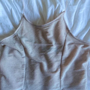 Glittrig beige linnetopp från Bik Bok - Säljer en glittrig beige linnetopp från Bik Bok med tunna axelband som man har runt nacken. Den har öppen rygg som gör den mer festlig.  Perfekt för att addera lite extra till din outfit. Tunt och lätt material som känns lyxigt mot huden. I storlek S