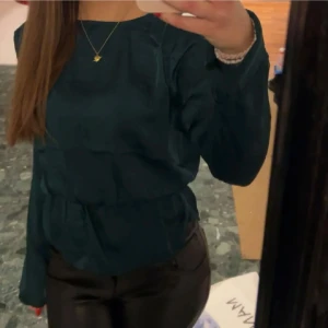 Mörkgrön omlottblus - Säljer en elegant mörkgrön omlottblus med långa ärmar och knytband i nacken. Blusen har en v-ringad design och en lätt volangdetalj vid midjan. Perfekt för en stilren look.