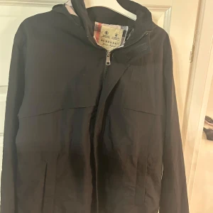 Burberry jacka - Säljer en stilren svart jacka från Burberry med klassiskt rutigt foder. Jackan har en dragkedja framtill och en huva med samma rutiga mönster. Perfekt för en elegant look.