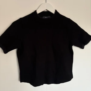 Svart stickad topp från Zara - Säljer en stilren svart stickad topp från Zara. Den har korta ärmar och en rund halsringning. Perfekt för en minimalistisk look. Passar bra till både jeans och kjol. Tröjan är i storlek M men passar mer som xs 