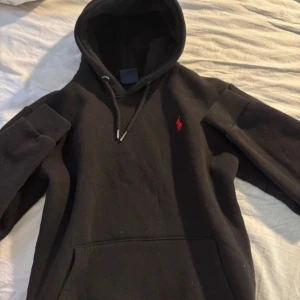Svart hoodie från Ralph Lauren - Säljer en klassisk svart hoodie från Ralph Lauren med röd logga på bröstet. Tröjan har en stor huva med dragsko och en praktisk magficka. Perfekt för en avslappnad stil.