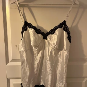 Vit satinlinne med spetsdetaljer - Elegant vit satinlinne med svarta spetsdetaljer längs kanterna. Axelbandslös design med justerbara axelband för perfekt passform. Perfekt för en chic och stilren look. Har tyvärr ett litet hål som syns på bilden. Men borde va lätt att sy ihop.