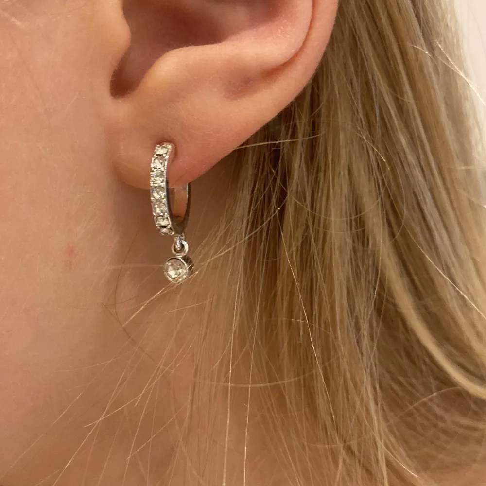 Snygga silverfärgade örhängen med små glittriga stenar och en hängande detalj. Perfekta för att ge lite extra bling till din outfit. De är små och diskreta men ändå iögonfallande.. Asusteet.