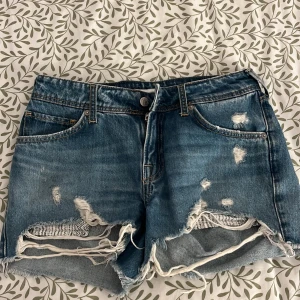 Blå jeansshorts med slitningar - Säljer ett par coola blå jeansshorts med slitningar. De har en klassisk femficksdesign och knappgylf. Perfekta för en avslappnad stil.
