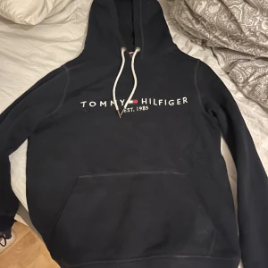 Svart hoodie från Tommy Hilfiger - Säljer en svart hoodie från Tommy Hilfiger med vit logga och snören. Perfekt för en casual look. Den har en klassisk känguruficka framtill och en bekväm passform.