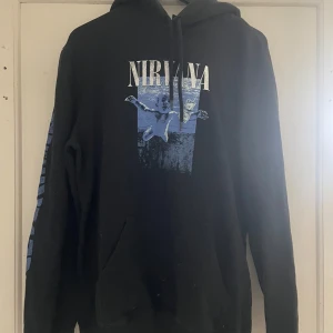 Svart Nirvana hoodie från H&M - Säljer en svart hoodie från H&M med Nirvana-tryck i blått och vitt. Tröjan har en klassisk hoodie-design med dragsko i huvan och en stor ficka framtill. Perfekt för fans av bandet! 🎸