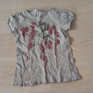 Grå t-shirt med blommönster - Säljer en grå t-shirt med ett rosa blommönster framtill. T-shirten har korta ärmar och är tillverkad i ett mjukt material. Perfekt för en avslappnad stil.