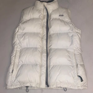 Vit dunväst från HVM - Snygg vit dunväst från HVM med dragkedja framtill. Västen har en hög krage och två praktiska fickor med dragkedja. Perfekt för kyligare dagar när du vill hålla värmen utan att kompromissa med stilen.