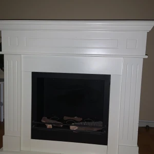 Vit eldstad i klassisk design spisbränsle  - Elegant vit eldstad med klassisk design och raka linjer. Den har en svart insats och dekorativa detaljer på framsidan. Perfekt för att skapa en mysig atmosfär i hemmet. Spisbränsle 