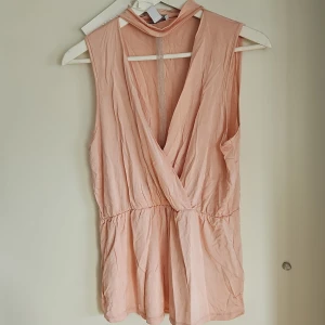 Rosa ärmlös omlottopp - Säljer en söt rosa ärmlös omlottopp med djup v-ringning och dekorativt band med pärlor i urringningen. Perfekt för varma dagar eller en kväll ute. Toppen har en luftig passform och är gjord i ett lätt material.