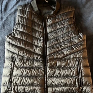 Grå quiltad dunväst - Snygg grå quiltad dunväst med dragkedja framtill. Västen har en stilren design med diagonala sömmar och praktiska fickor med dragkedja. Perfekt för lager-på-lager under kyligare dagar.