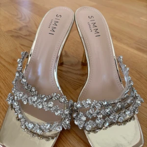 Guldiga klackskor från Simmi London - Helt nya, snygga guldiga klackskor från Simmi London med glittriga detaljer. Skorna har en elegant design med öppen tå och smala remmar dekorerade med strass.  Storlek 41 men passar även 40!