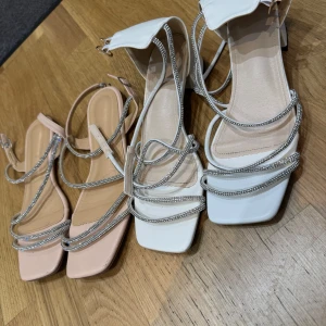 Eleganta sandaler med strassdetaljer - Snygga sandaler i beige och vit med glittriga strassdetaljer. De har en elegant design med flera tunna remmar och en liten klack. Perfekta för att ge en touch av glamour till din outfit. Endast använda 1 gång. 200kr för båda. Strl 42.