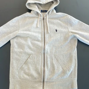 Grå hoodie från Polo Ralph Lauren - Säljer en stilren grå hoodie från Polo Ralph Lauren med dragkedja och broderad logga på bröstet. Tröjan har en klassisk design med huva och fickor framtill. Perfekt för en avslappnad look.