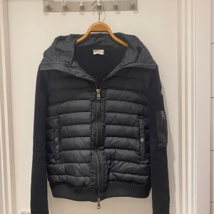 Svart jacka från Moncler - Säljer en snygg svart jacka från Moncler med quiltad design och dragkedja framtill. Jackan har långa ärmar och en huva för extra skydd. Perfekt för kyligare dagar med sin stilrena look och Moncler-logga på ärmen.