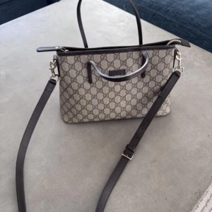 Gucciväska med axelrem - Snygg handväska med klassiskt mönster och avtagbar axelrem. Väskan har en stilren design med mörka handtag och detaljer. Perfekt för att ge din outfit en lyxig touch.