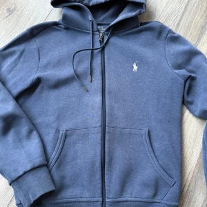 Blå hoodie från Ralph Lauren - Säljer en snygg blå hoodie från Ralph Lauren med dragkedja och klassisk logga på bröstet. Huvan har justerbara snören för extra komfort. Snygg blå färg nu till våren och sommaren! 