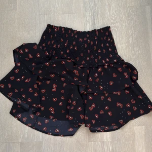 mörk blå kjol med röds blommor  - Mörk blå kjol med röda blommor och volangdetaljer. Den har en elastisk midja för en bekväm passform. Perfekt för en söt och lekfull stil.