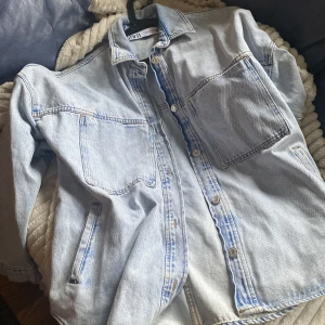Ljusblå jeansjacka från ZARA - Säljer en stilren ljusblå jeansjacka från ZARA. Jackan har en klassisk krage och knäppning framtill med metallknappar. Den har två bröstfickor och två sidofickor, perfekt för en casual look. Passar perfekt för vår och sommar!