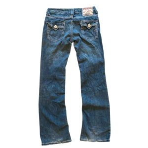 True Religion jeans bootcut lågmidjade - Snygga blå jeans från True Religion med klassisk femficksdesign och dekorativa sömmar. Byxorna är bootcut och lågmidjade en tydlig logotyp på baksidan. Perfekta för en avslappnad stil. Ytterben 100 cm, innerben 75 cm, midja 37cm. Bra skick men lite heeldrag