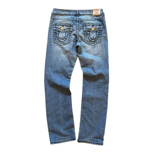 True Religion jeans  - Snygga blå jeans från True Religion med unika bakfickor och slitna detaljer. De har en klassisk femficksdesign och knappar med märkets logga. Perfekta för en avslappnad stil. Ytterben 102 cm, innerben 79 cm, midja 39 cm