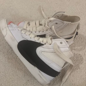 Vita Nike Blazer sneakers - Säljer ett par vita Nike Blazer sneakers med svart swoosh. Skorna har hög modell med snörning och en klassisk design. Perfekta för en stilren look. Köpte för jag ville ha de till gymmet men tyvärr lite för små för mig! 