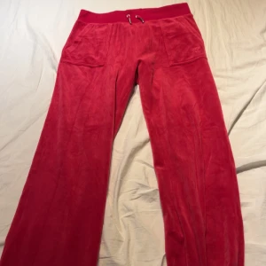 Röda mjukisbyxor från Juicy Couture - Snygga röda mjukisbyxor från Juicy Couture med broderad JC-logga. Byxorna har en loose passform och är tillverkade i mjukt material. De har snörning i midjan och fickor både fram och bak med knappar.