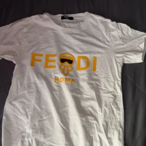 Vit t-shirt från Fendi - Snygg vit t-shirt från Fendi med ett gult tryck framtill som inkluderar texten 'FEODI ROMA' och en stiliserad figur. Perfekt för en avslappnad stil med en touch av lyx.