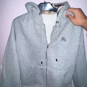 Grå kofta från Burberry - Säljer en stilren grå huvtröja från Burberry med klassiskt rutigt mönster på insidan av huvan. Tröjan har dragkedja framtill och praktiska fickor. Perfekt för en avslappnad look med en touch av lyx. Den e storlek L men passar även M 