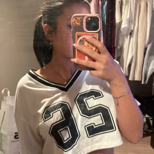 Vit croppad topp med nummertryck - Säljer en vit croppad topp med svart nummertryck '26' på framsidan. Toppen har en v-ringad hals med svarta ränder. Perfekt för en sportig look!