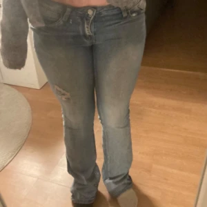 Blå jeans med slitningar - Snygga blå jeans med en avslappnad passform och slitningar på ena benet. Perfekta för en casual look. De har en klassisk femficksdesign och stängs med knapp och dragkedja.