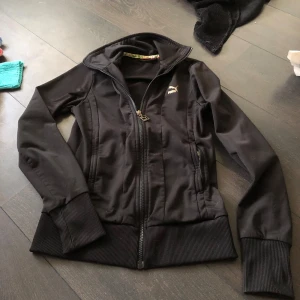 Svart jacka från Puma - Snygg svart jacka från Puma med dragkedja och ribbade muddar. Jackan har en hög krage och två fickor med dragkedja. Perfekt för en sportig look. Storlek XS-S skulle jag tro? 