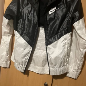 Svart och vit vindjacka från Nike - Snygg svart och vit vindjacka från Nike med dragkedja och huva. Jackan har en sportig look med långärmade ärmar och elastiska muddar. Perfekt för blåsiga dagar.