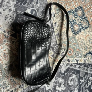 Svart axelväska från NLY - Snygg svart axelväska från NLY med krokodilpräglat mönster. Väskan har en dragkedja upptill och en justerbar axelrem. Perfekt för att ge en stilren touch till din outfit. (Pruta på!)