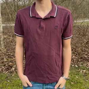 Vinröd pikétröja från Fred Perry - Säljer en snygg vinröd pikétröja från Fred Perry med klassisk krage och knappar. Tröjan har vita och blå ränder på krage och ärmslut, vilket ger en stilren look. Perfekt för en avslappnad stil.