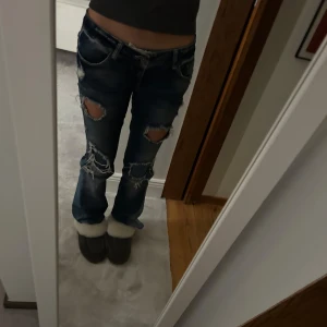 Blå slitna jeans - Säljer ett par blå jeans med slitningar och hål på knäna. De har en låg midja och en bootcut-stil. Perfekta för en avslappnad look. Midjemått: 39cm❤️ innerbenslängd: 77cm! OBS: är omsydda nere för att få bootcut, och en cm i grenen, men inget som syns alls.