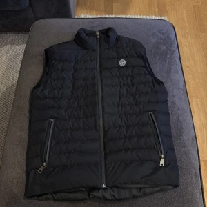 Svart dunväst från Stone Island - Säljer en stilren svart dunväst från Stone Island. Västen har en dragkedja framtill och två praktiska fickor med dragkedjor. Perfekt för kyligare dagar och ger en snygg look med sin quiltade design.