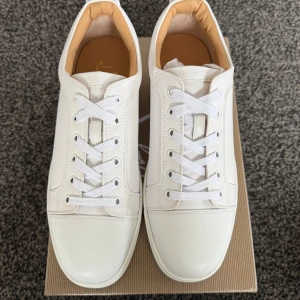 Vita sneakers från Christian Louboutin - Snygga vita sneakers från Christian Louboutin med den ikoniska röda sulan. Skorna har snörning och är tillverkade i skinn, vilket ger en lyxig känsla. Perfekta för den som vill ha stilrena och eleganta skor.