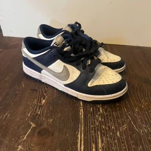 Nike Dunk i blått och vitt - Snygga Nike Dunk sneakers i en klassisk blå och vit färgkombination. Skorna har en stilren design med snörning och den ikoniska Nike-loggan på sidan. Perfekta för en casual look.
