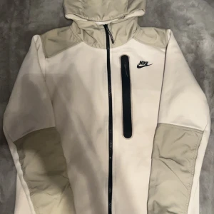 Nike tech tröja  - Säljer en stilren vit och beige jacka från Nike med huva och dragkedja. Jackan har en svart Nike-logga på bröstet och en vertikal dragkedja på framsidan. Perfekt för en sportig look!