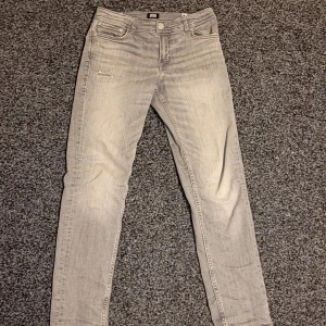 Grå jeans från Jack & Jones - Snygga grå jeans från Jack & Jones med en slim passform. De har en klassisk femficksdesign och en diskret slitning på framsidan. Perfekta för en stilren look.
