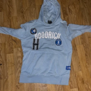 Ljusblå hoodie från Hoodrich män och kvinna - Säljer en ljusblå hoodie från Hoodrich med coola patches och textdetaljer. Hoodien har en stor ficka framtill och en bekväm huva. Perfekt för en avslappnad stil.