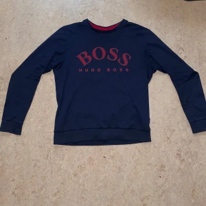 Hugo Boss Tröja - Säljer en snygg mörkblå tröja från Hugo Boss med röd logga på framsidan. Tröjan har långa ärmar och en rund halsringning med röd detalj på insidan. Tröjan har även en QR kod på sidan. Om man har flera frågor så är det bara till att skriva. 