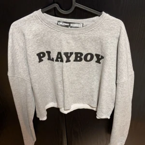 Grå croppad tröja från Playboy - Snygg grå croppad tröja från Playboy med svart text på framsidan. Tröjan har långa ärmar och en avslappnad passform. Perfekt för en casual look.