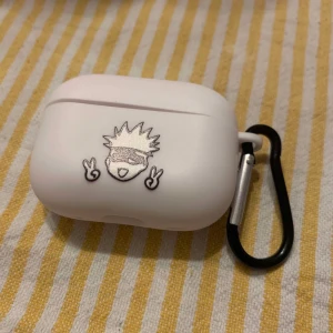 Vit AirPods-fodral med karbinhake - Säljer ett vitt AirPods-fodral med en cool tecknad figur på framsidan. Fodralet har en praktisk svart karbinhake för enkel fastsättning på väskan eller nyckelringen. Perfekt för att skydda dina AirPods med stil!